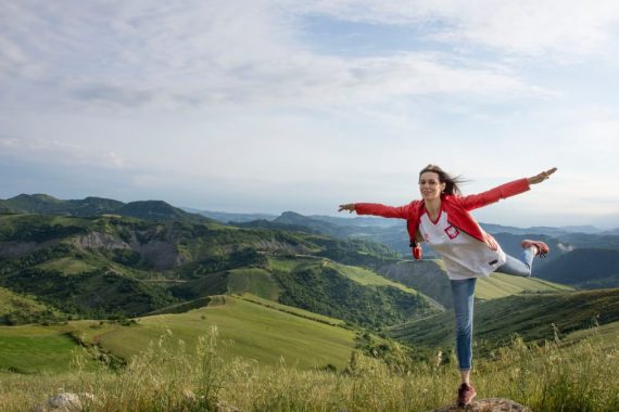 A woman joyfully balances on a rock amidst rolling green hills under a clear blue sky.
